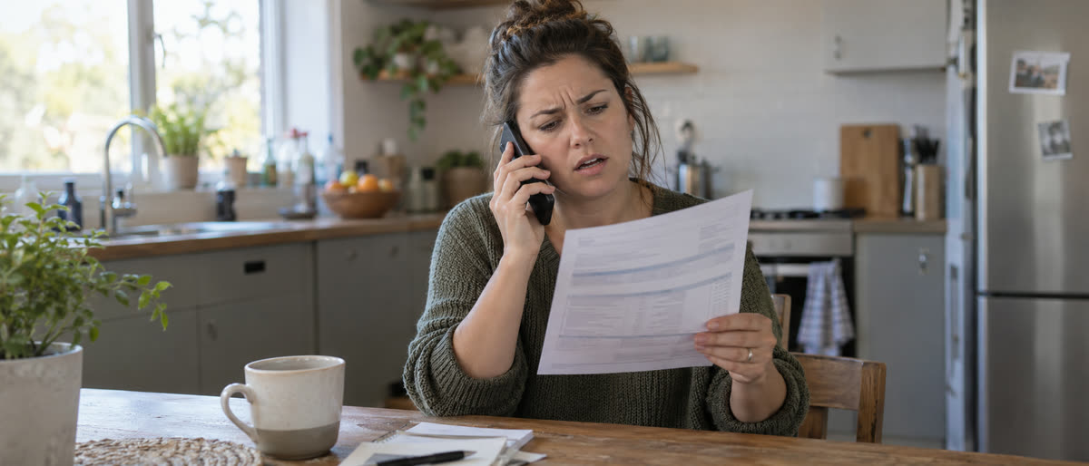 An exasperated customer on a phone call holds up a printed bill at a kitchen table.